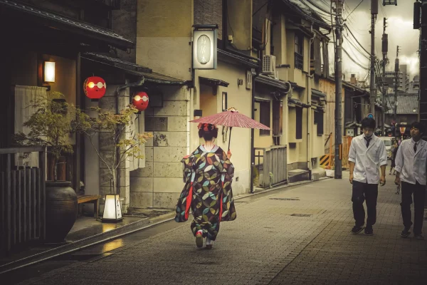 Une geisha japonaise tenant un parapluie rouge descend une rue de Gion à Kyoto tandis que deux hommes passent sans oser la regarder