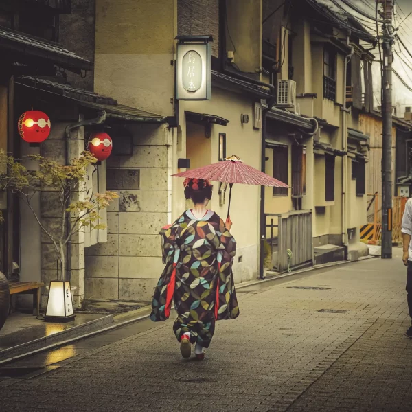 Une geisha japonaise tenant un parapluie rouge descend une rue de Gion à Kyoto tandis que deux hommes passent sans oser la regarder