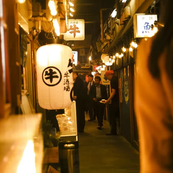 Woman walking in a somewhat crowded narrow alley lit by all the lanterns and lights of the multiple restaurants boarding it left and right.