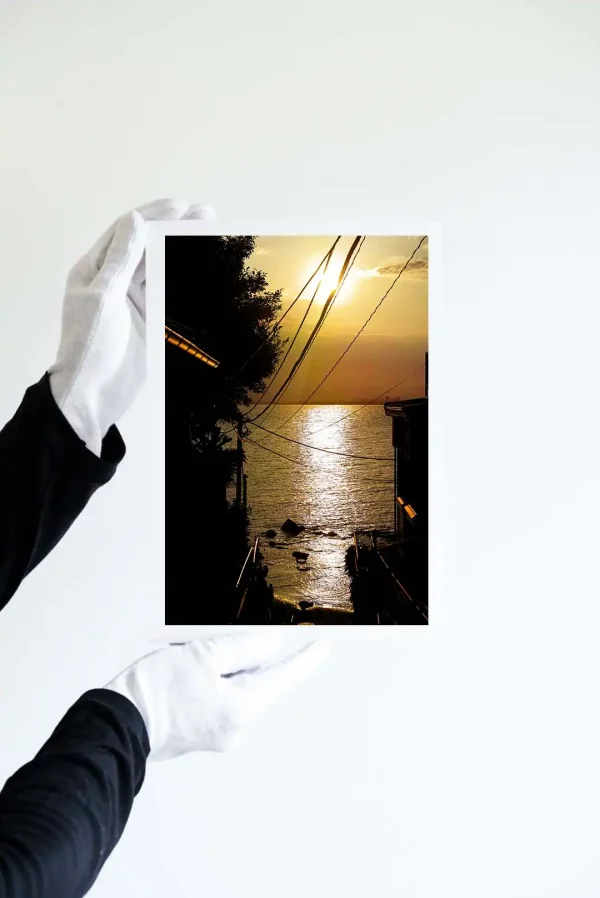 Hands in white gloves holding a fine art print of The Sagami bay viewed from the top of stairs from Enoshima Island during a beautiful and blazing sunset.